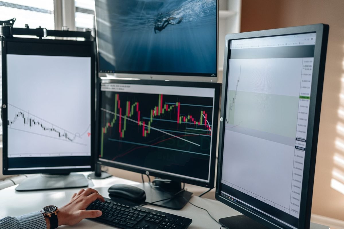 Desk with three monitors showing charts and a person using a keyboard