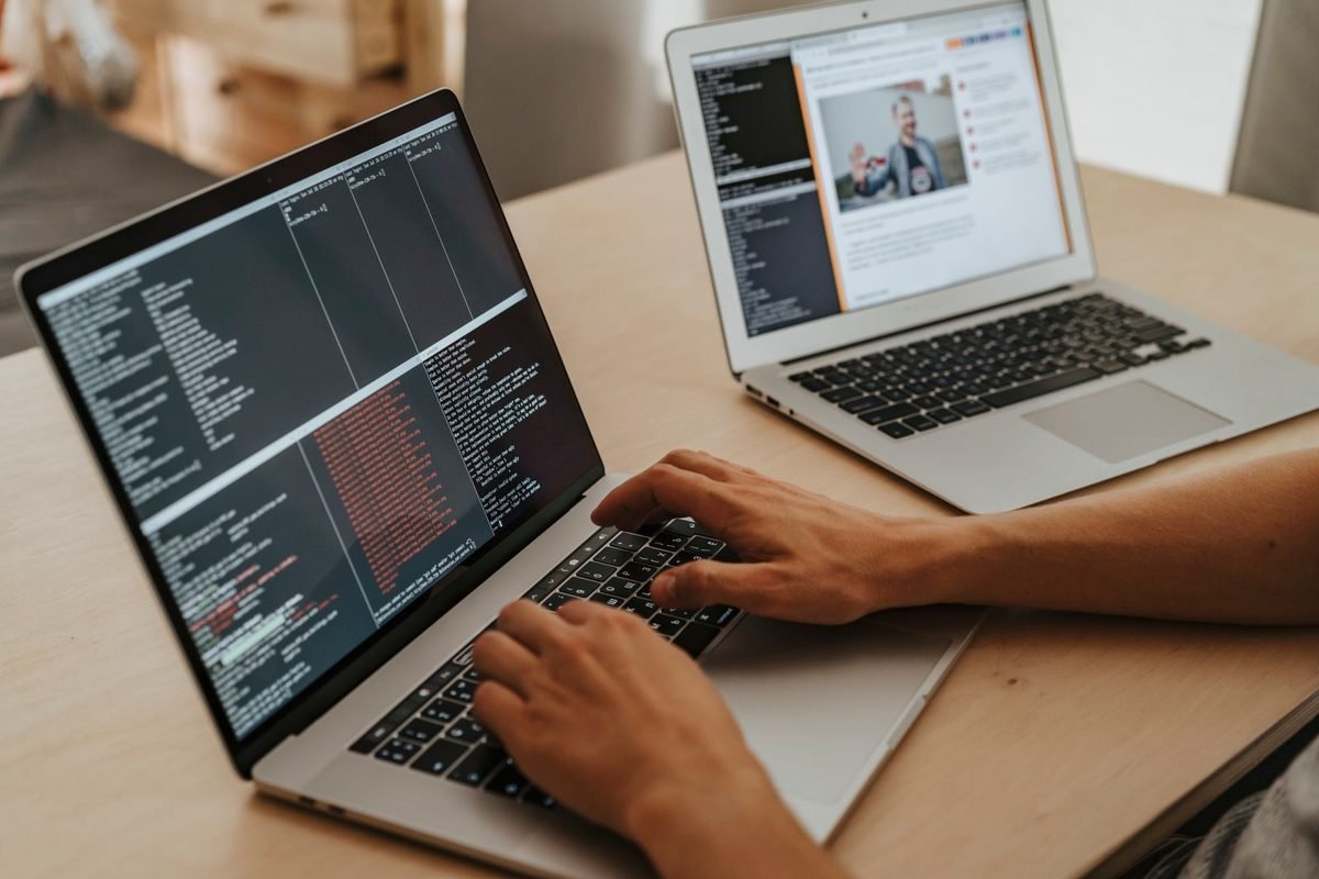 Hands typing on a laptop showing lines of code next to a second laptop